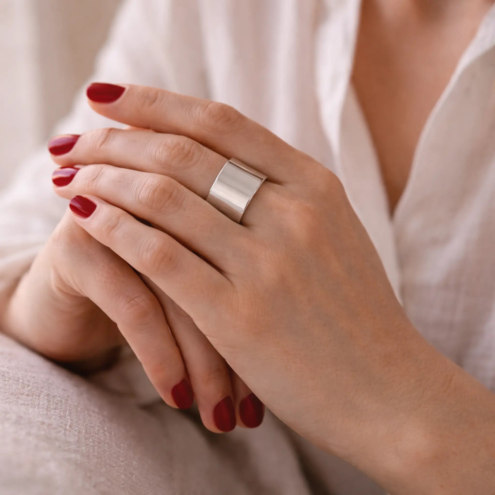Nahaufnahme: Hände mit rot lackierten Nägeln vor weißer Bluse – breiter, glänzender Silber-Bandring am Finger, weiches Licht.