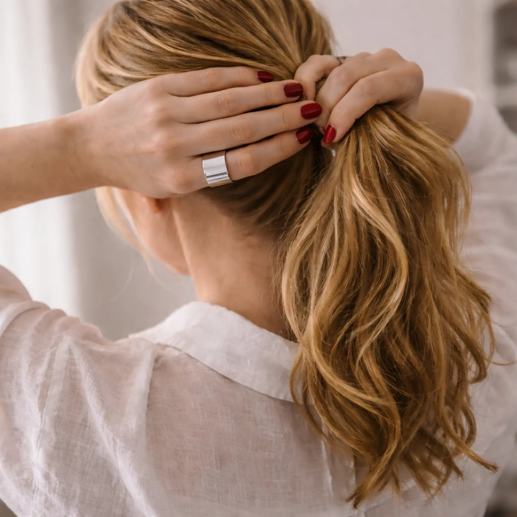 Rückansicht: Frau bindet einen Pferdeschwanz – rot lackierte Nägel und breiter, glänzender Silber-Bandring am Finger, weiße Leinenbluse.
