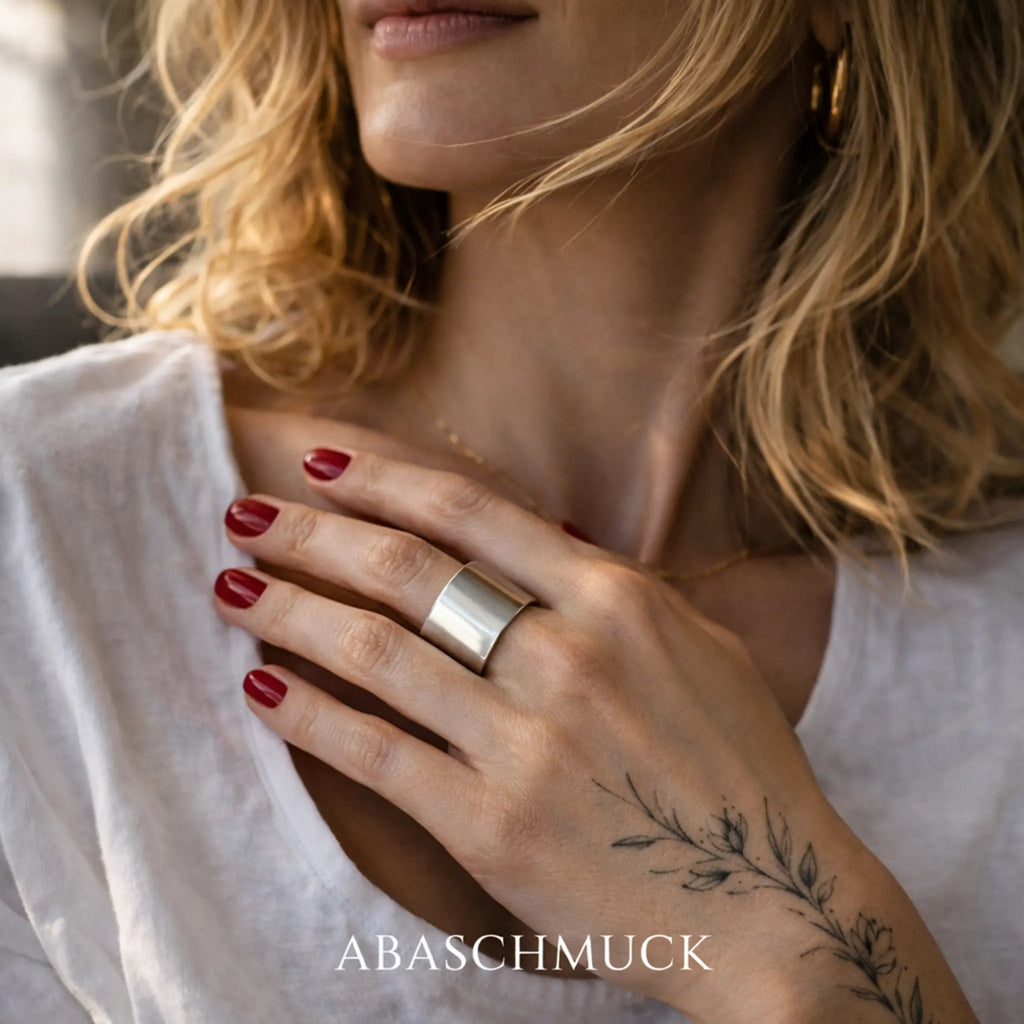 Nahaufnahme: Blondine in weißer Bluse mit rot lackierten Nägeln hält die Hand auf die Brust – breiter, glänzender Silber-Bandring am Finger, zartes Zweig-Tattoo am Handgelenk.