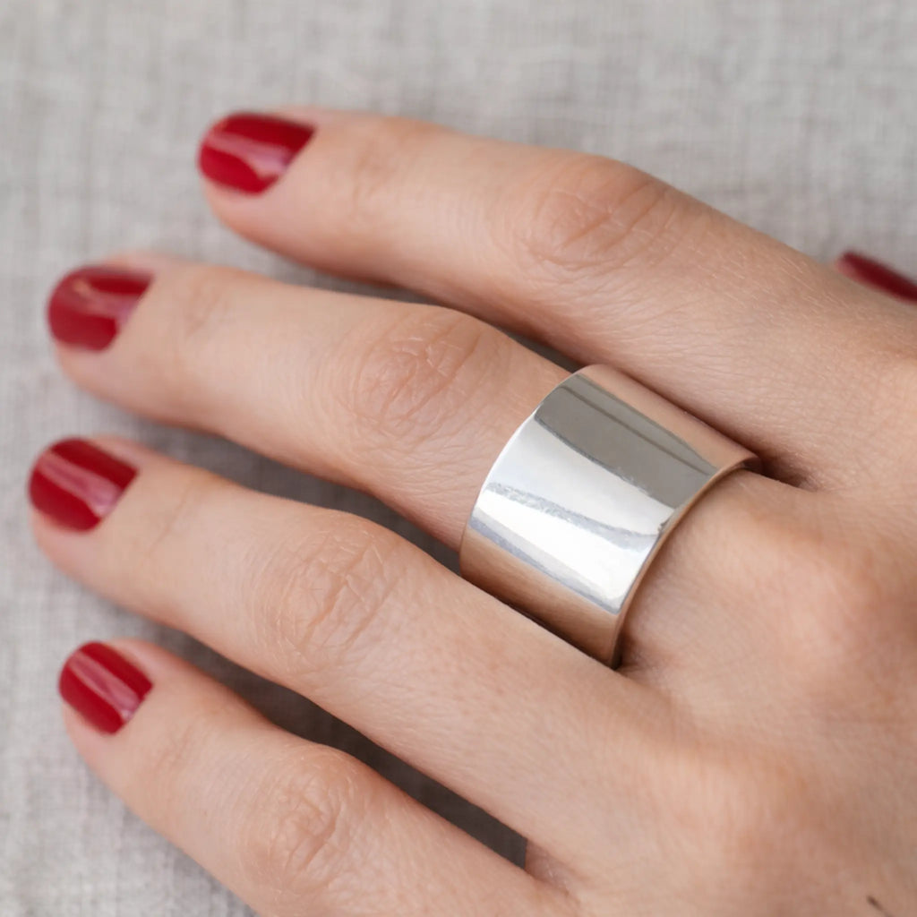 Detailaufnahme einer Hand mit rot lackierten Nägeln – breiter, glänzender Silberring (Bandring) am Finger vor hellem Leinenhintergrund.