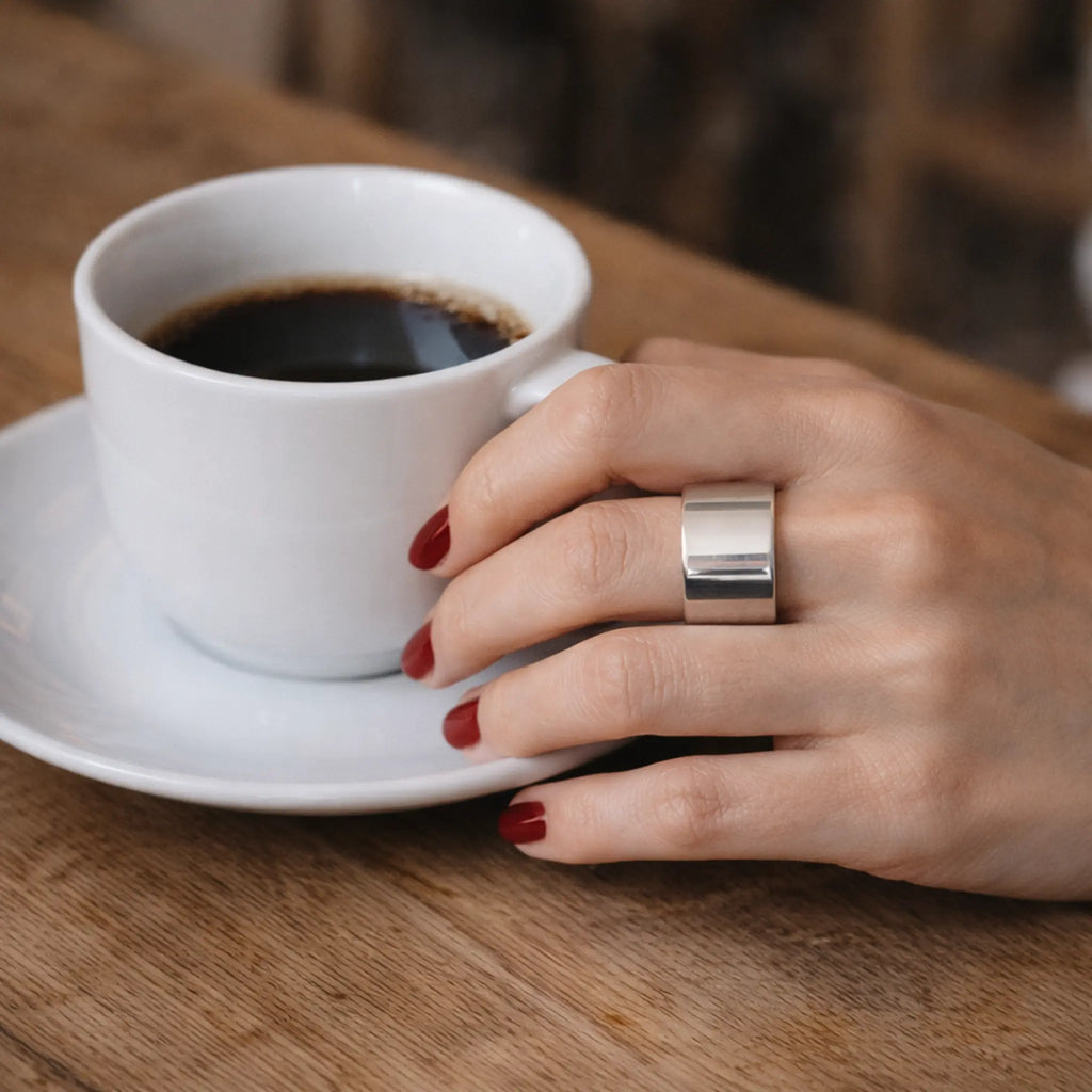Hand mit rot lackierten Nägeln hält eine weiße Kaffeetasse – breiter, glänzender Silber-Bandring am Finger auf Holztisch.