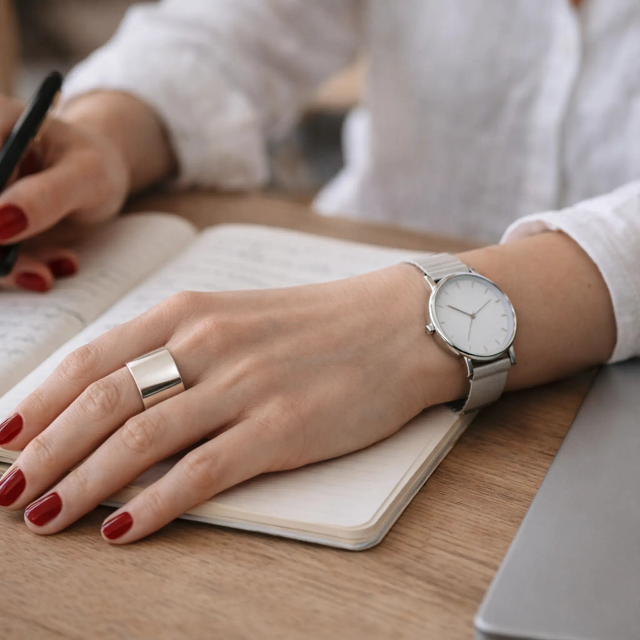 Hand mit rot lackierten Nägeln schreibt in ein Notizbuch – breiter, glänzender Silber-Bandring am Finger, silberne Armbanduhr, Holztisch im Hintergrund.