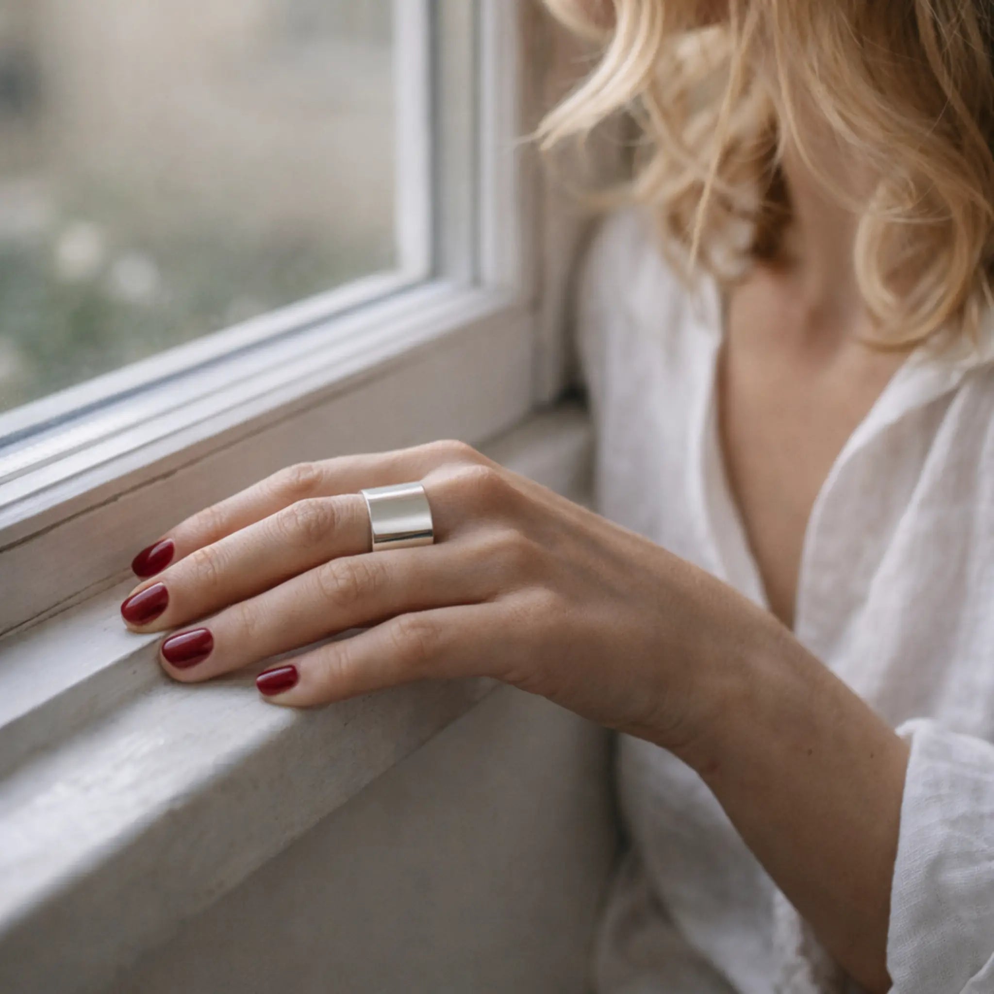 Blondine in weißer Leinenbluse am Fenster – Hand mit rot lackierten Nägeln auf der Fensterbank, breiter glänzender Silber-Bandring am Finger.