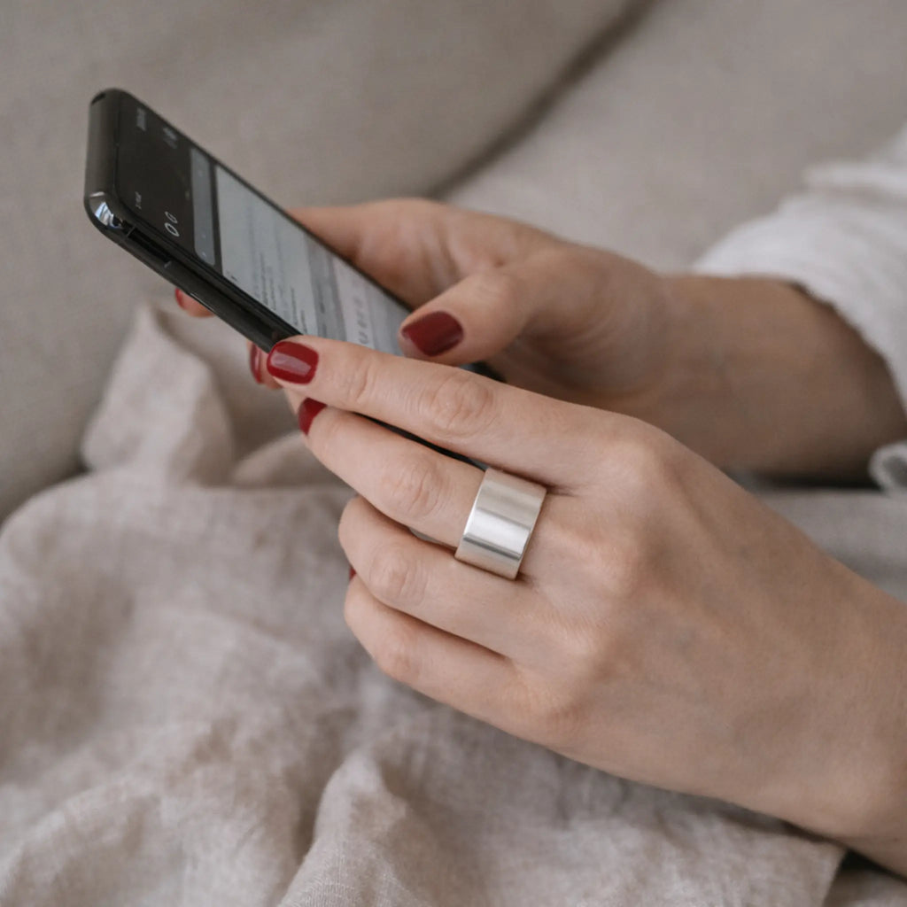 Nahaufnahme: Hände mit rot lackierten Nägeln halten ein Smartphone – breiter, glänzender Silber-Bandring am Finger, entspannte Szene auf hellem Stoff/Sofa.