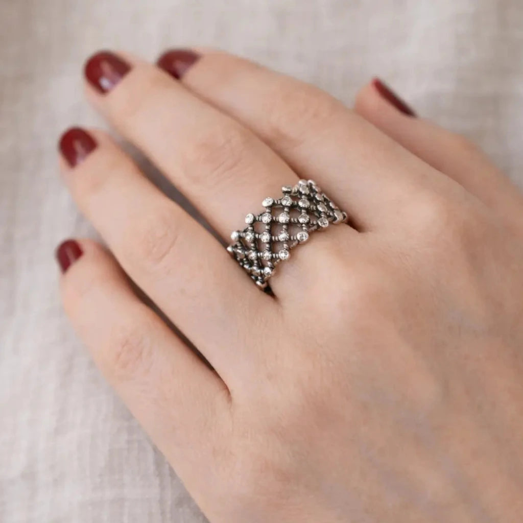 Nahaufnahme einer Hand mit bordeauxroten Nägeln; am Finger ein breiter, filigraner Silberring mit netzartigem Muster und kleinen Kugeln, heller Stoffhintergrund.