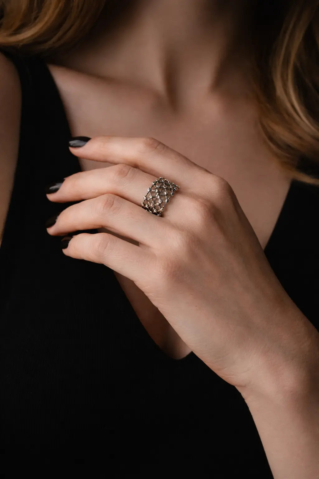Nahaufnahme einer Hand mit dunkel lackierten Nägeln vor schwarzem Kleid; am Finger ein filigraner silberner Ring mit netzartigem Muster, elegantes Licht.