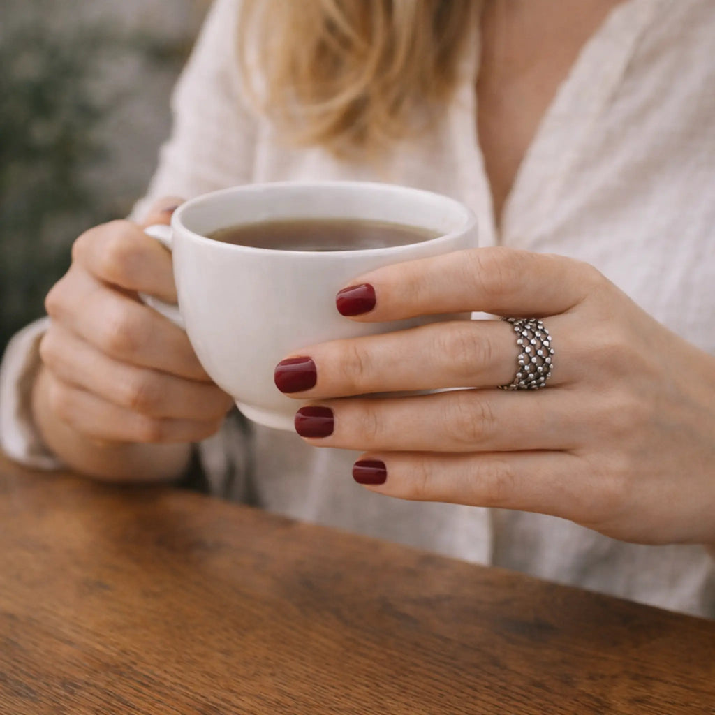 Nahaufnahme: Hände mit bordeauxroten Nägeln halten eine weiße Kaffeetasse; am Finger ein breiter, filigraner Silberring mit netzartigem Muster, helle Bluse und Holztisch im Hintergrund.
