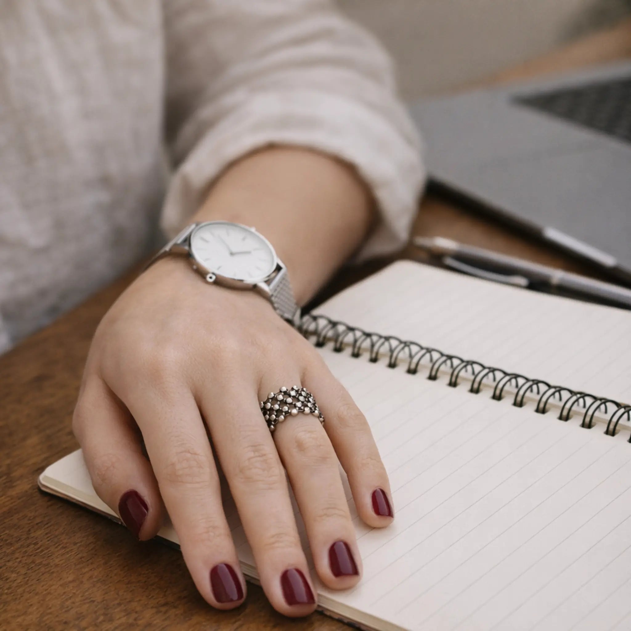 Hand mit bordeauxroten Nägeln liegt auf einem Notizbuch; silberner Ring mit netzartigem Muster und Armbanduhr sichtbar, Schreibtisch mit Laptop im Hintergrund.