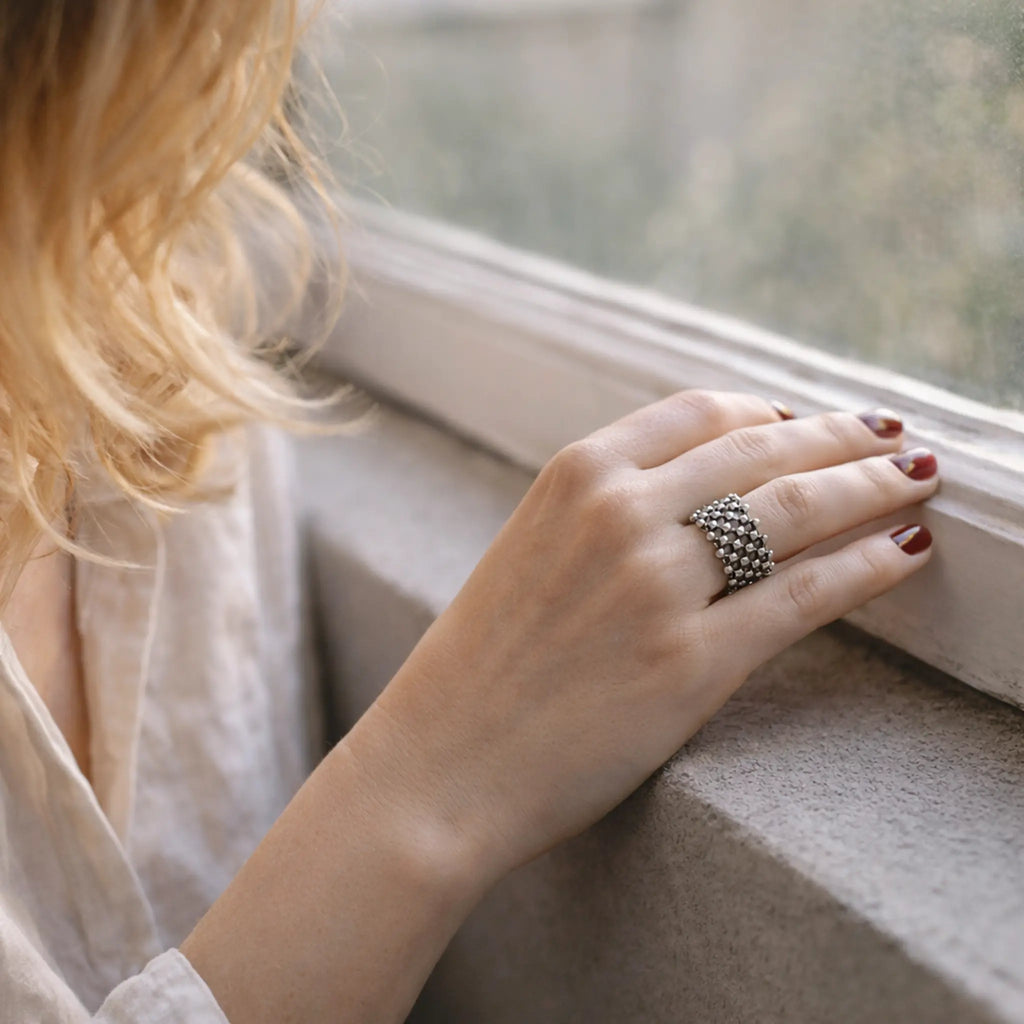 Hand einer Frau am Fensterbrett mit bordeauxroten Nägeln; trägt einen breiten, filigranen Silberring mit netzartigem Muster, weiches Tageslicht, helle Bluse und blonde Haare im Hintergrund.