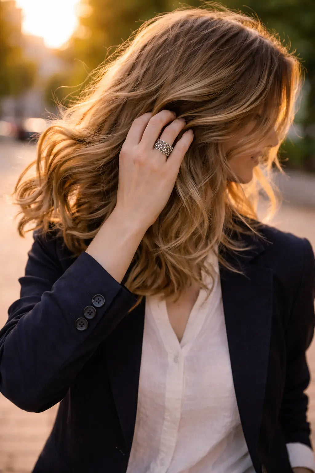 Frau im Blazer im warmen Abendlicht, Hand im Haar; am Finger ein filigraner silberner Ring mit netzartigem Muster, Hintergrund in weichem Bokeh.