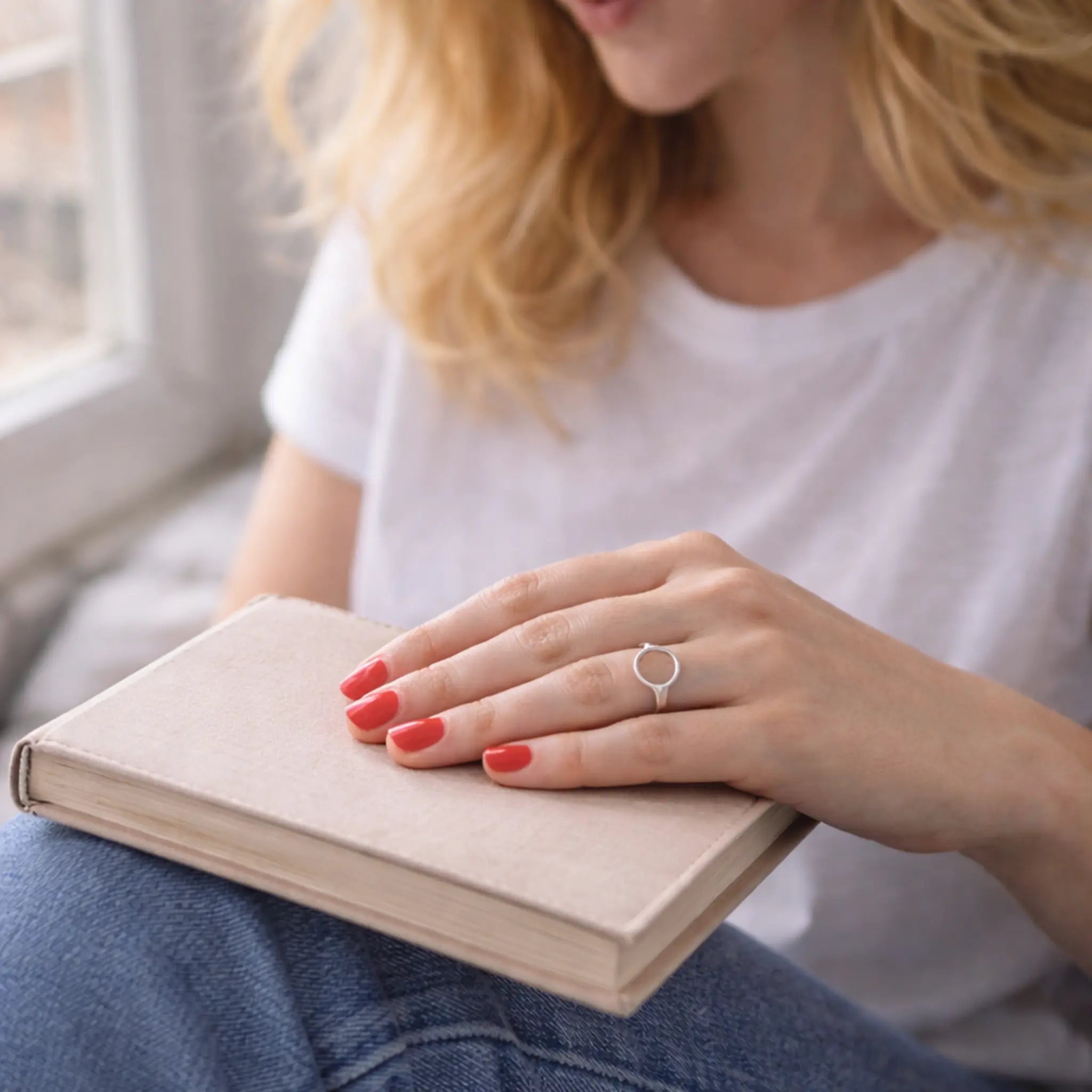 Hand auf geschlossenem Buch, roter Nagellack, minimalistischer Silberring als Kreis – moderner Damenring, clean und zeitlos.