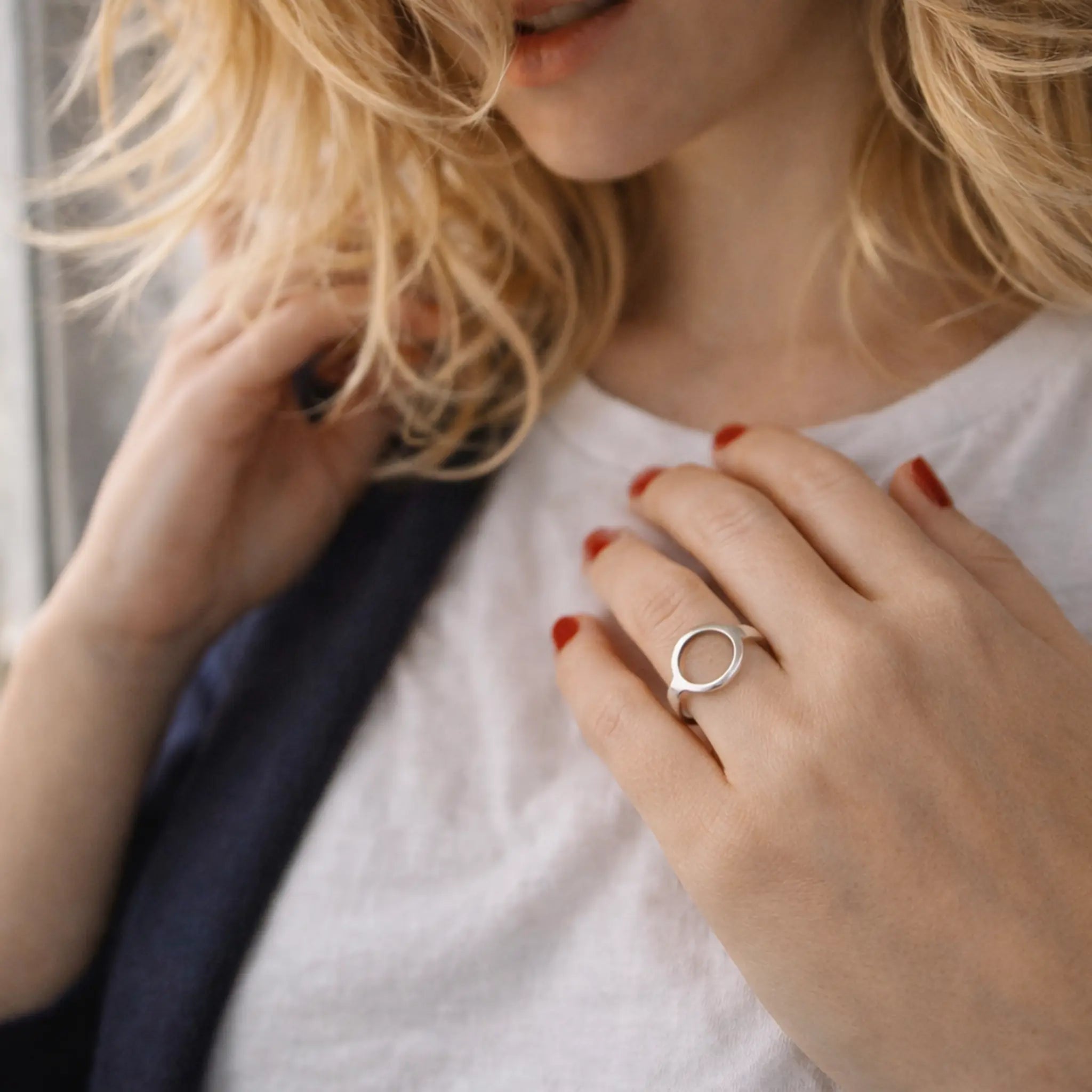 Blonde Frau am Fenster, Hand am Dekolleté mit minimalistischem Kreis-Silberring – eleganter Damenring, rote Nägel, weiches Tageslicht.