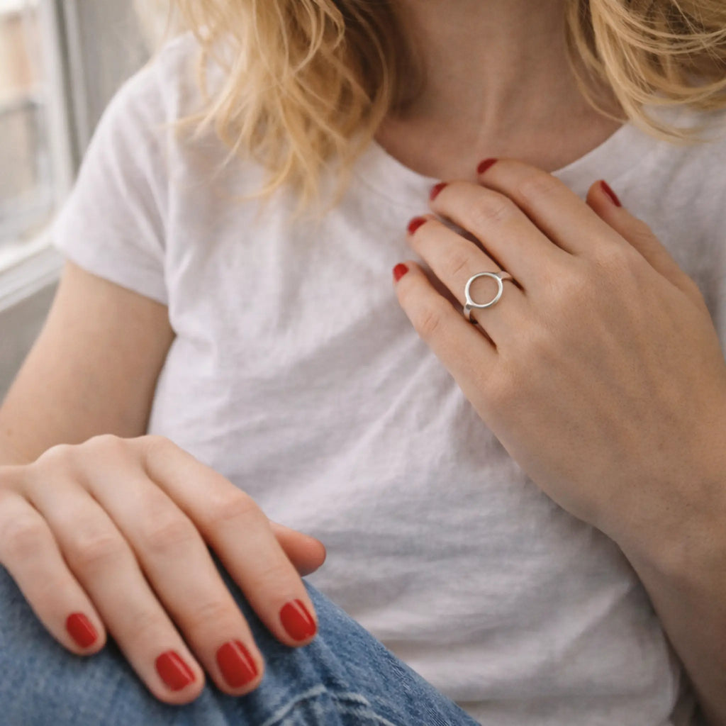 Frau im weißen T-Shirt und Jeans am Fenster, roter Nagellack, minimalistischer Silberring im Kreis-Design – dezenter Alltags-Damenring.