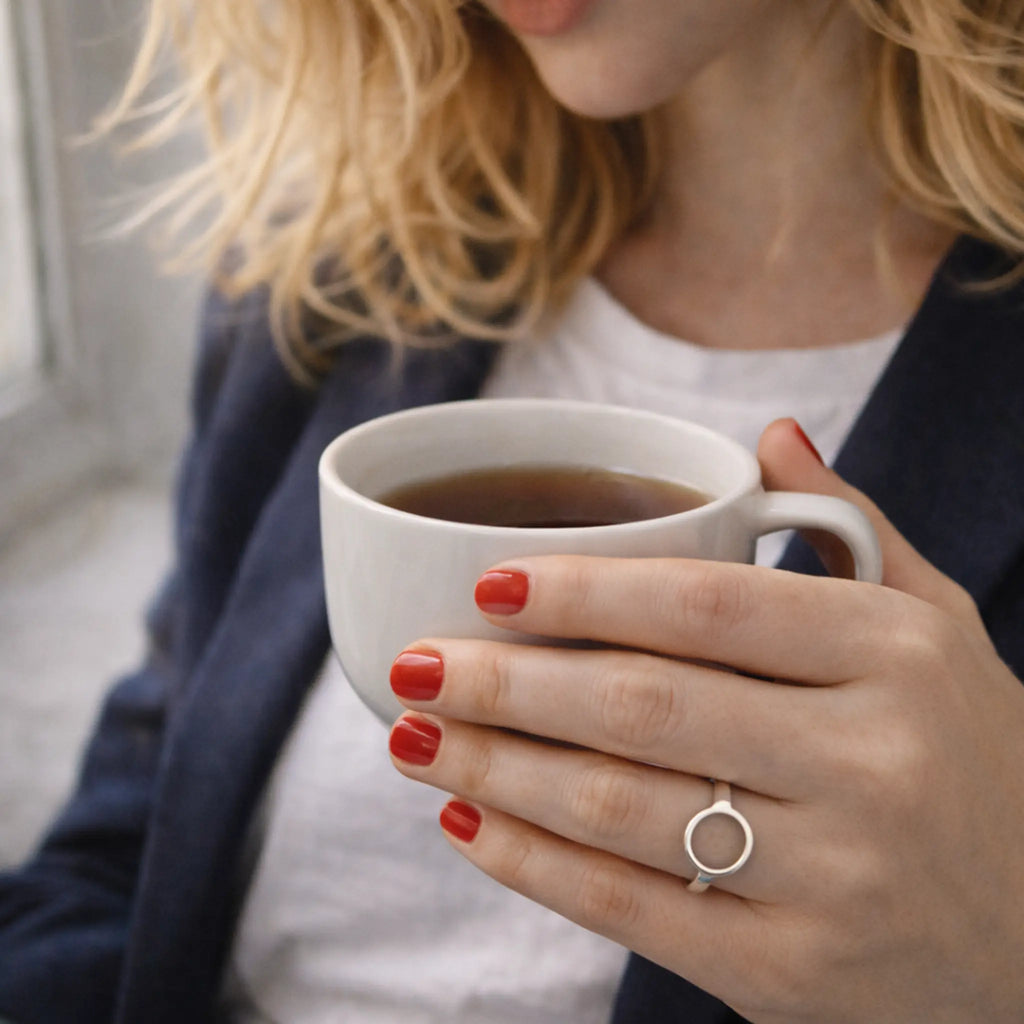 Hand mit rotem Nagellack hält Kaffeetasse, minimalistischer Silberring mit Kreis-Motiv – zeitloser Damenring, Cozy Lifestyle am Fenster.