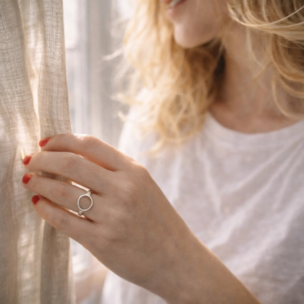 Hand mit Kreis-Silberring zieht einen leichten Vorhang zur Seite, rote Nägel – minimalistischer Damenring, natürliche Lichtstimmung.