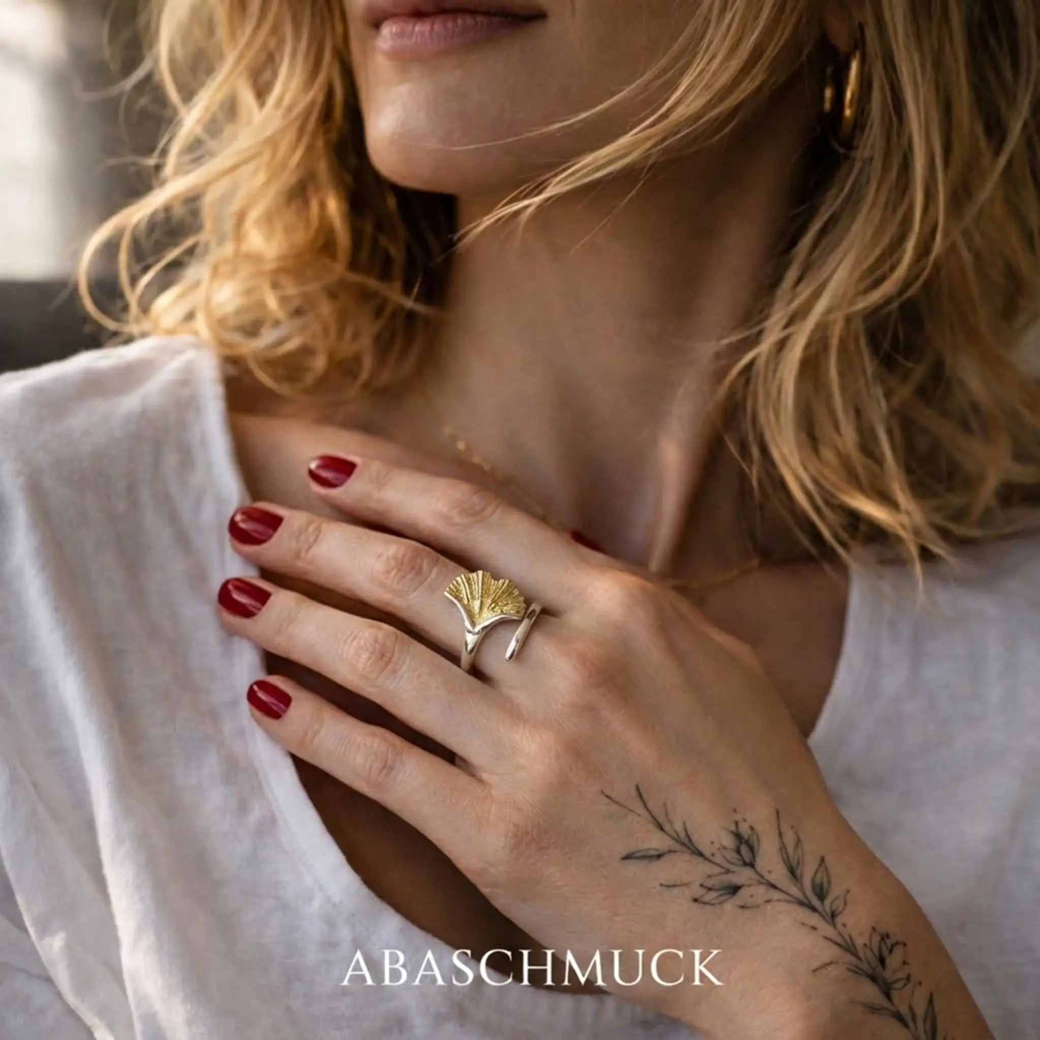 Blonde Frau in weißer Bluse, Hand am Dekolleté mit Silberring und vergoldetem Ginkgo-Blatt – eleganter Damenring im Natur-Design, warmes Licht.