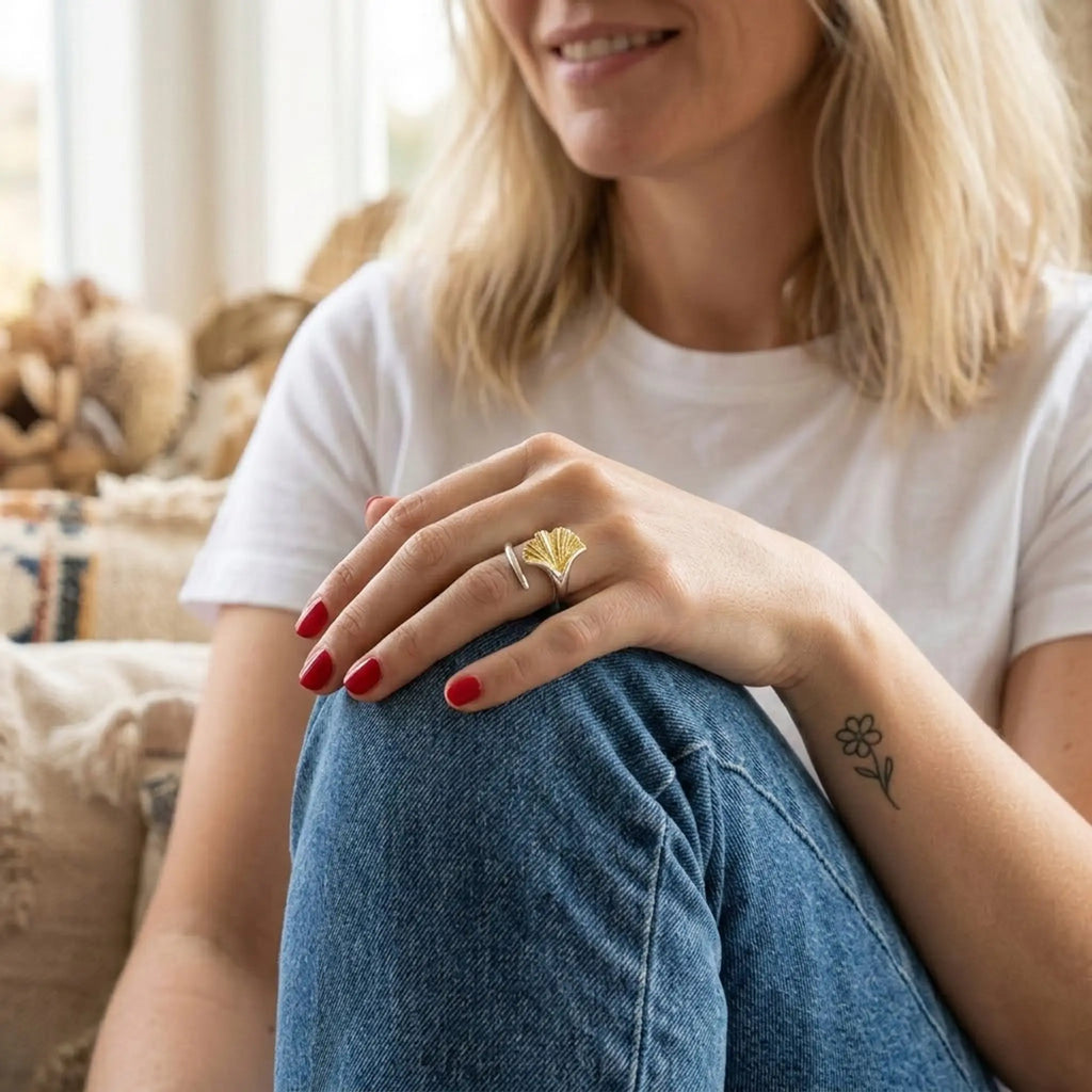 Frau in weißem T-Shirt und Jeans sitzt entspannt; Silberring mit vergoldetem Ginkgo-Blatt am Finger – lässiger Alltagsschmuck, Lifestyle-Foto.