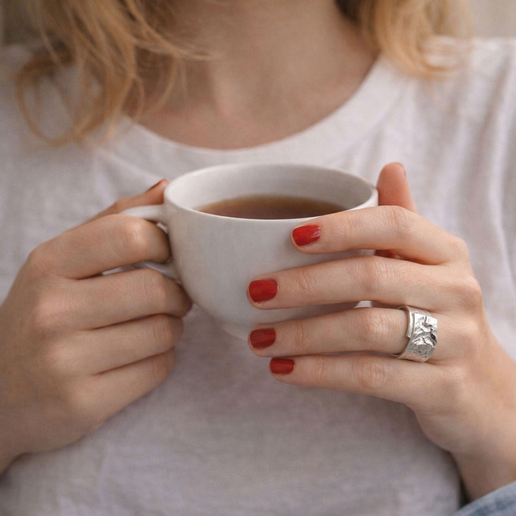 Frau mit rotem Nagellack hält eine weiße Tasse in den Händen, breiter strukturierter 925 Sterling Silberring als eleganter Statement Ring für Damen im Fokus