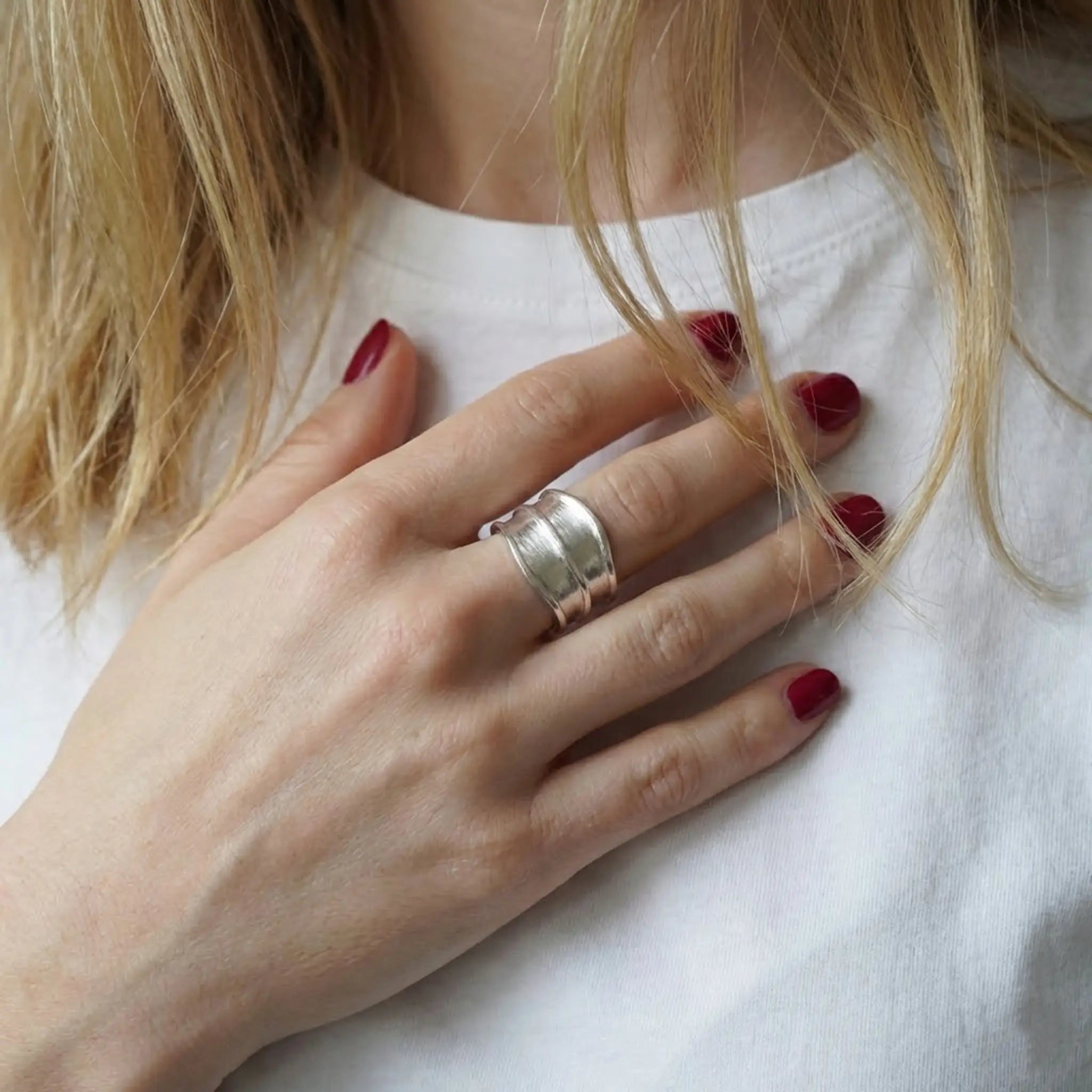 Nahaufnahme: Hand mit bordeauxroten Nägeln auf der Brust, breiter Damenring aus Silber mit matter Oberfläche, weißes T-Shirt.