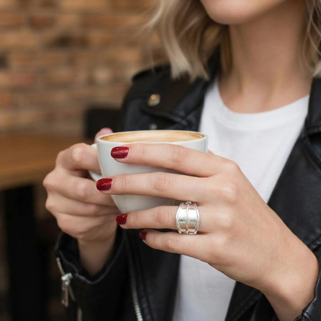 Frau hält eine Tasse Kaffee; rote Nägel und breiter Silberring am Finger, weißes Shirt und Lederjacke, Café-Lifestyle.