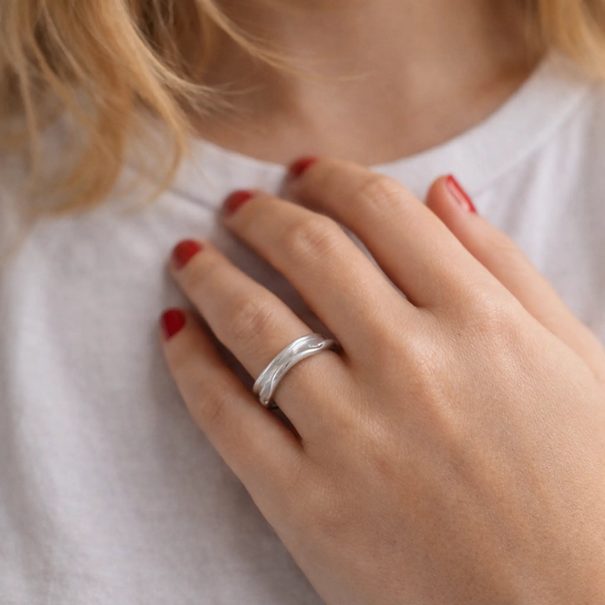 Nahaufnahme: Hand mit roten Nägeln auf der Brust, minimalistischer Damenring aus Silber mit sanft gewellter Struktur, weißes T-Shirt, Lifestyle-Foto.