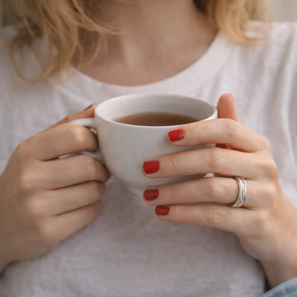 Frau hält eine weiße Tasse Tee/Kaffee; rote Nägel und minimalistischer Damenring aus Silber am Finger, weißes T-Shirt, gemütliches Lifestyle-Motiv.
