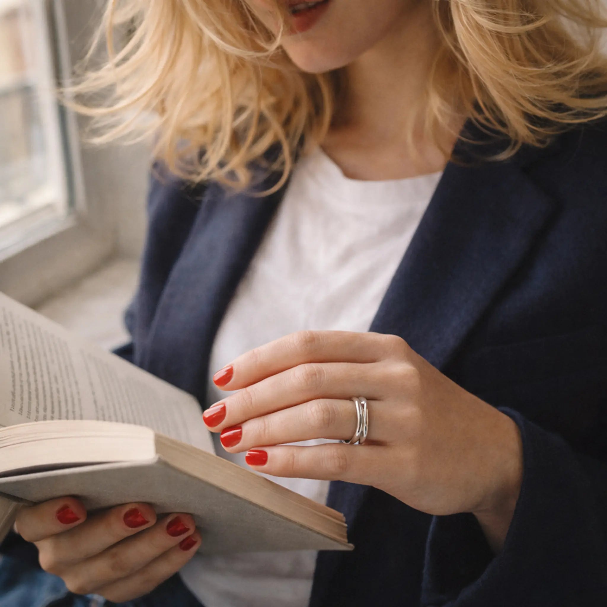 Frau liest ein Buch am Fenster, trägt weißen Shirt und dunklen Blazer; rote Nägel und minimalistischer Silberring am Finger, elegante Lifestyle-Aufnahme.
