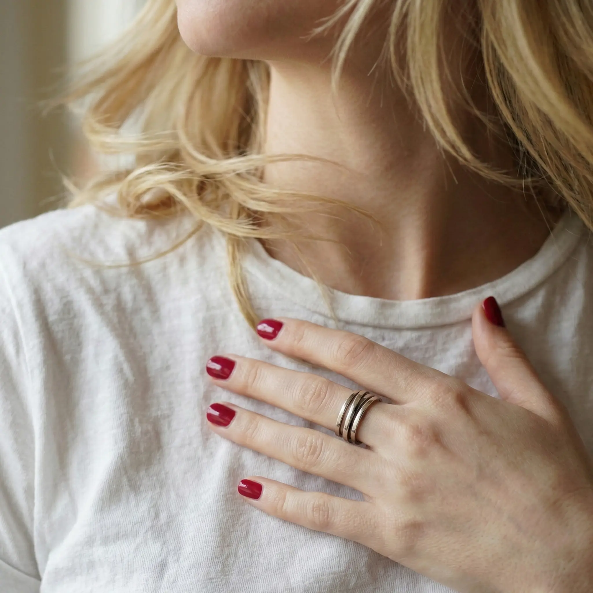 Blonde Frau mit leicht gewelltem Haar trägt einen modernen 925 Sterling Silberring mit drei schmalen parallelen Bändern. Die Hand mit rot lackierten Nägeln liegt elegant am Schlüsselbein, weißes Baumwollshirt und natürliches Licht betonen den minimalistischen Silberring als stilvollen Alltagsschmuck für Frauen.