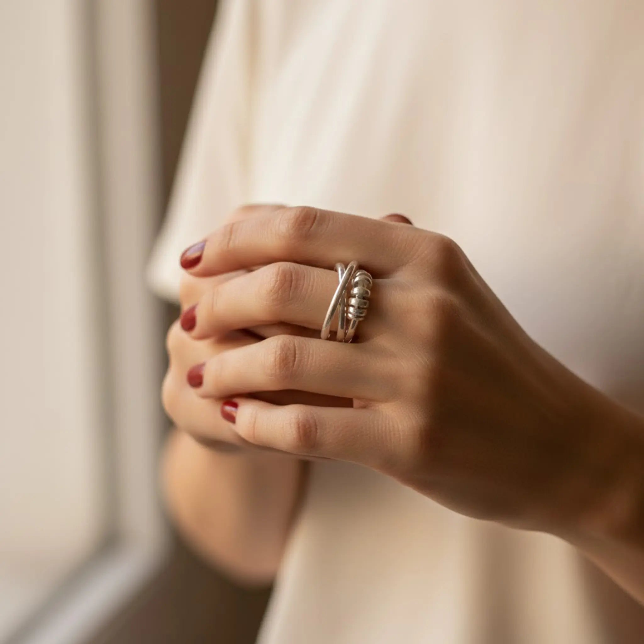 Nahaufnahme von ineinander verschränkten Händen mit kurzen rot lackierten Nägeln; am Finger ein silberner Ring mit Wickel-/Knoten-Design, warmes Tageslicht, weißes Shirt im Hintergrund.