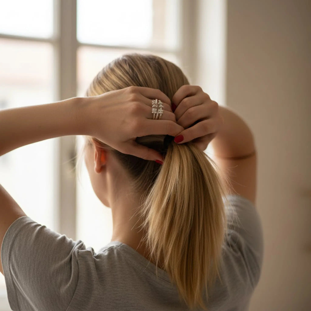 Blonde Frau von hinten bindet sich einen Pferdeschwanz; rote Nägel und silberner Ring am Finger sichtbar, graues Shirt, weiches Tageslicht am Fenster.