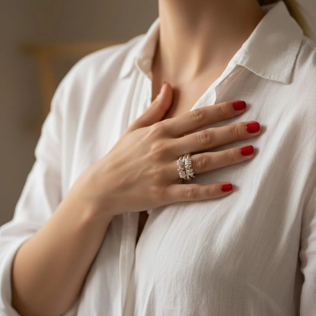 Nahaufnahme: Hand einer Frau auf der Brust über einer weißen Bluse, kurze rot lackierte Nägel; silberner Ring am Finger, weiches warmes Tageslicht.