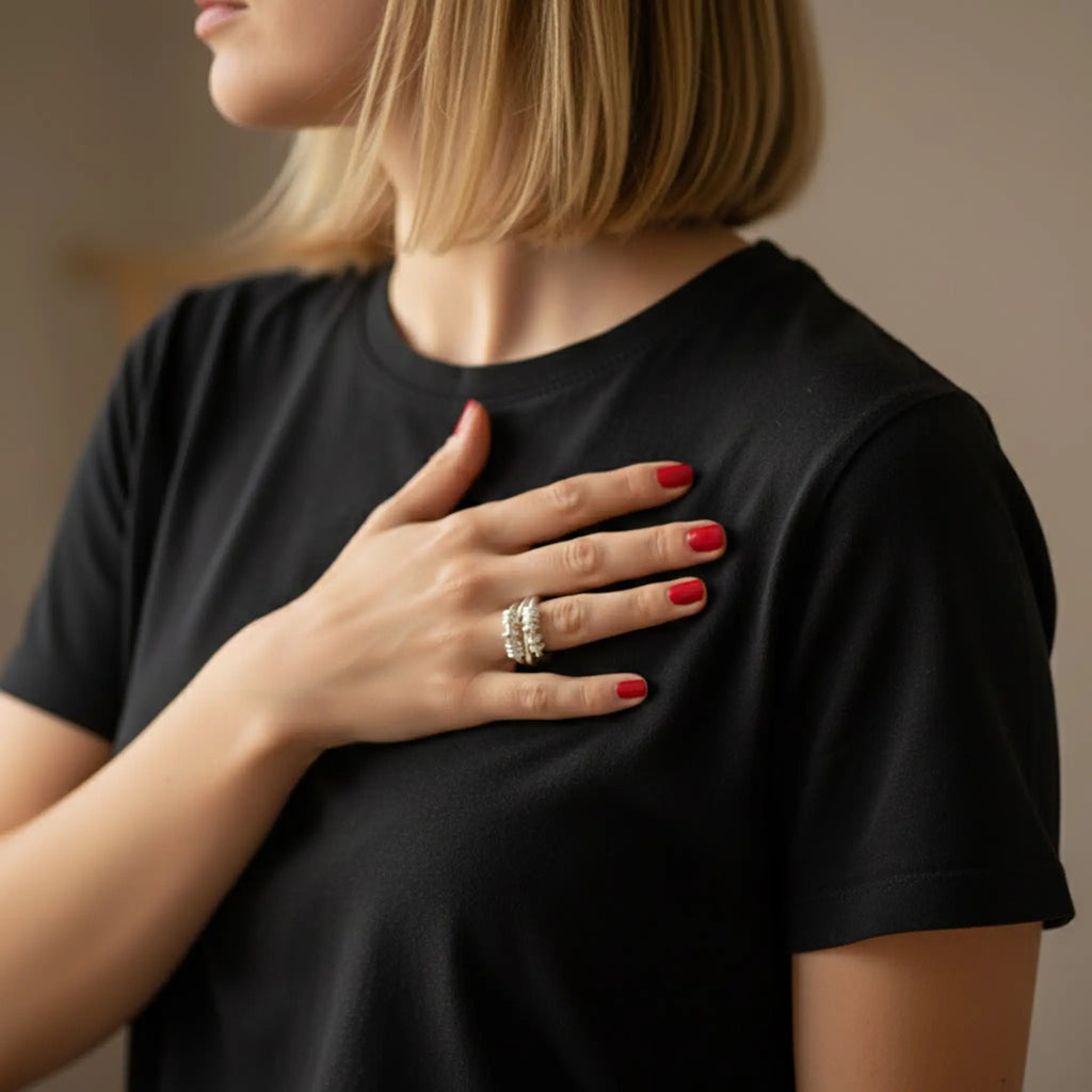Frau im schwarzen Shirt im Profil, Hand auf der Brust, kurze rot lackierte Nägel; silberner Ring am Finger, weiches warmes Licht, Hintergrund unscharf.