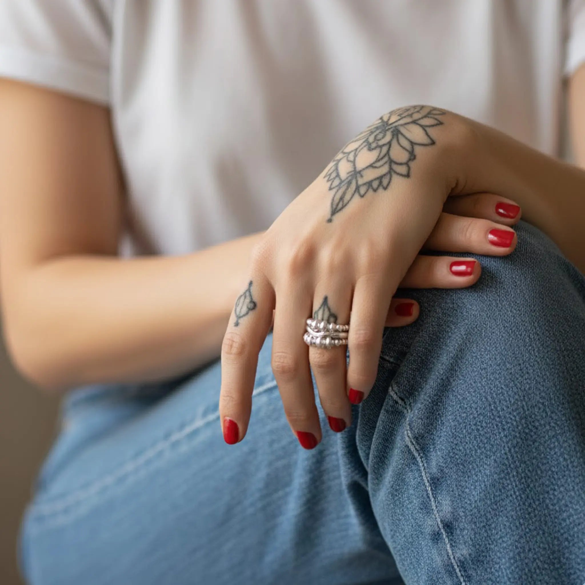 Nahaufnahme einer Hand mit floralem Tattoo und kurzen rot lackierten Nägeln auf Jeans; am Finger ein silberner Ring, weißes Shirt im Hintergrund, weiches Tageslicht.