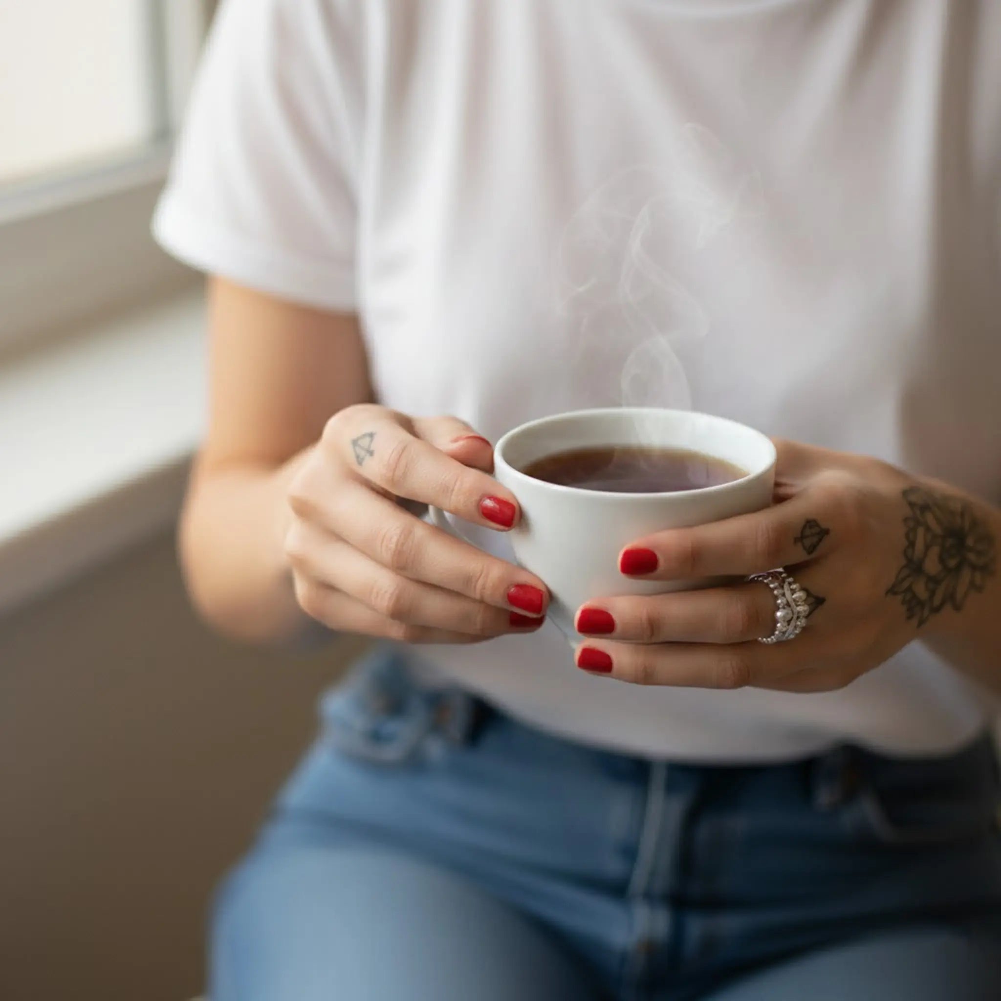 Frau in weißem T-Shirt und Jeans hält eine dampfende Tasse Kaffee; rote Nägel, Tattoos an den Händen und silberner Ring am Finger, weiches Tageslicht am Fenster.