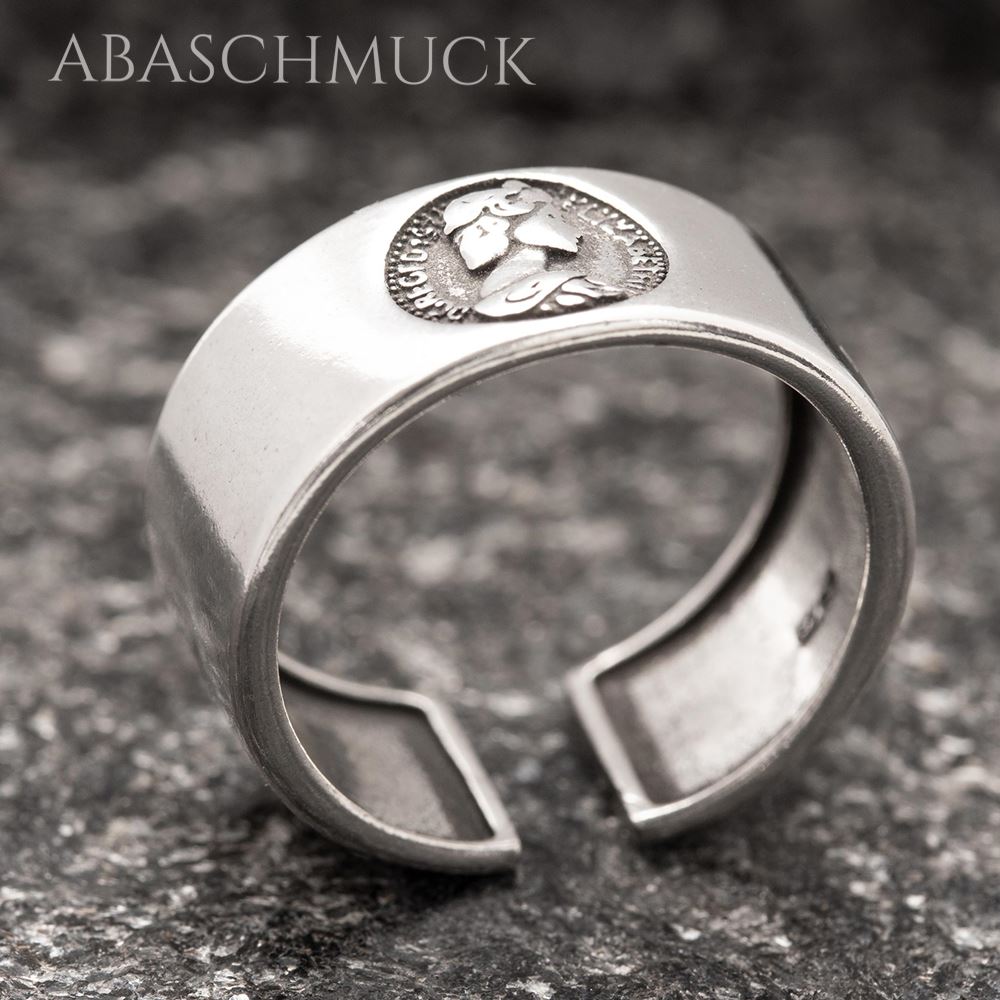 Schrägansicht eines modernen Silberrings mit rundem Münzmotiv auf dunkler Steinfläche.