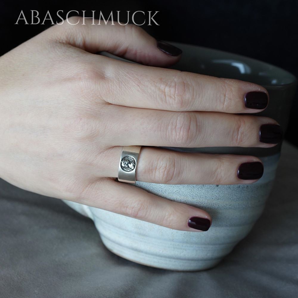 
Handaufnahme mit breitem Silberring und Münzemblem, dunkler Nagellack.
