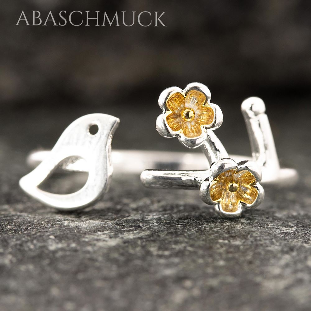 Silberner, verstellbarer Ring mit Vogelmotiv und goldenen Blüten aus der Nähe, liegend auf grauer Oberfläche.