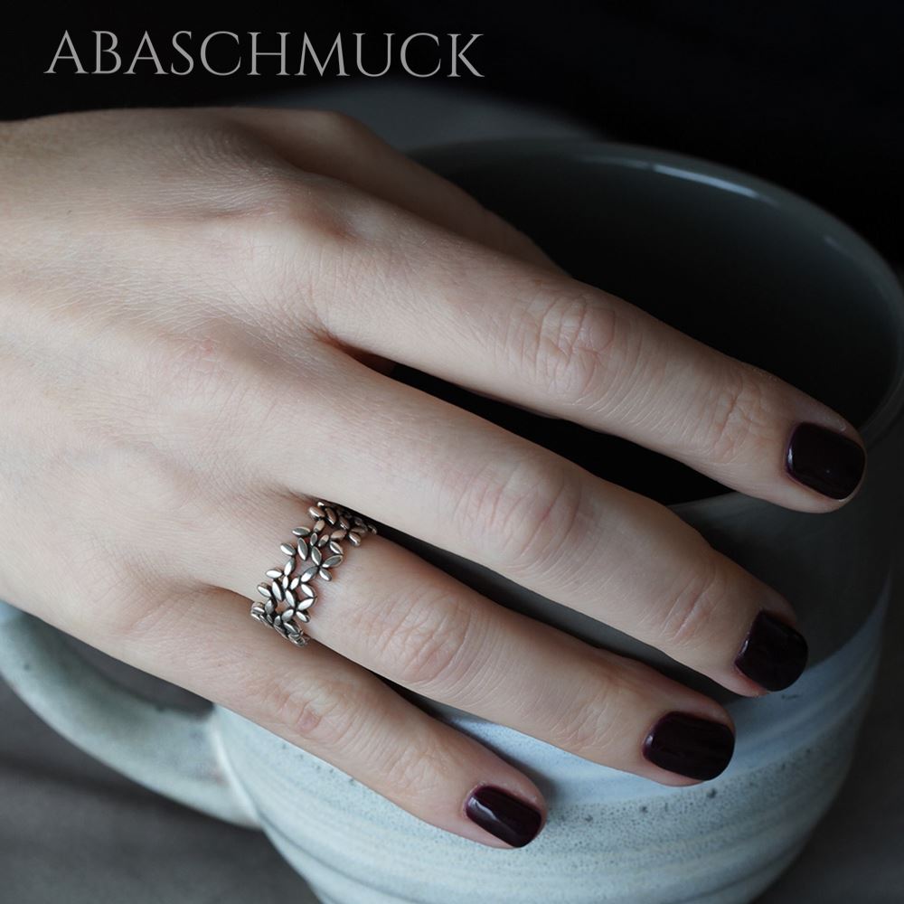 Hand mit dunkel lackierten Nägeln trägt einen offenen Silberring mit filigranem Blätterdesign und hält eine Keramiktasse.