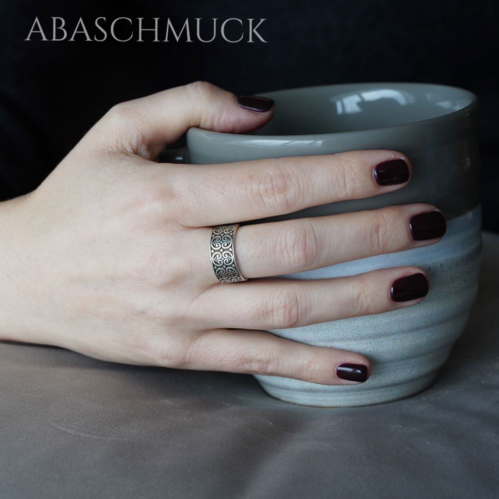Hand mit dunkel lackierten Nägeln trägt einen breiten Silberring mit Ornamentmuster und hält eine graue Keramiktasse.