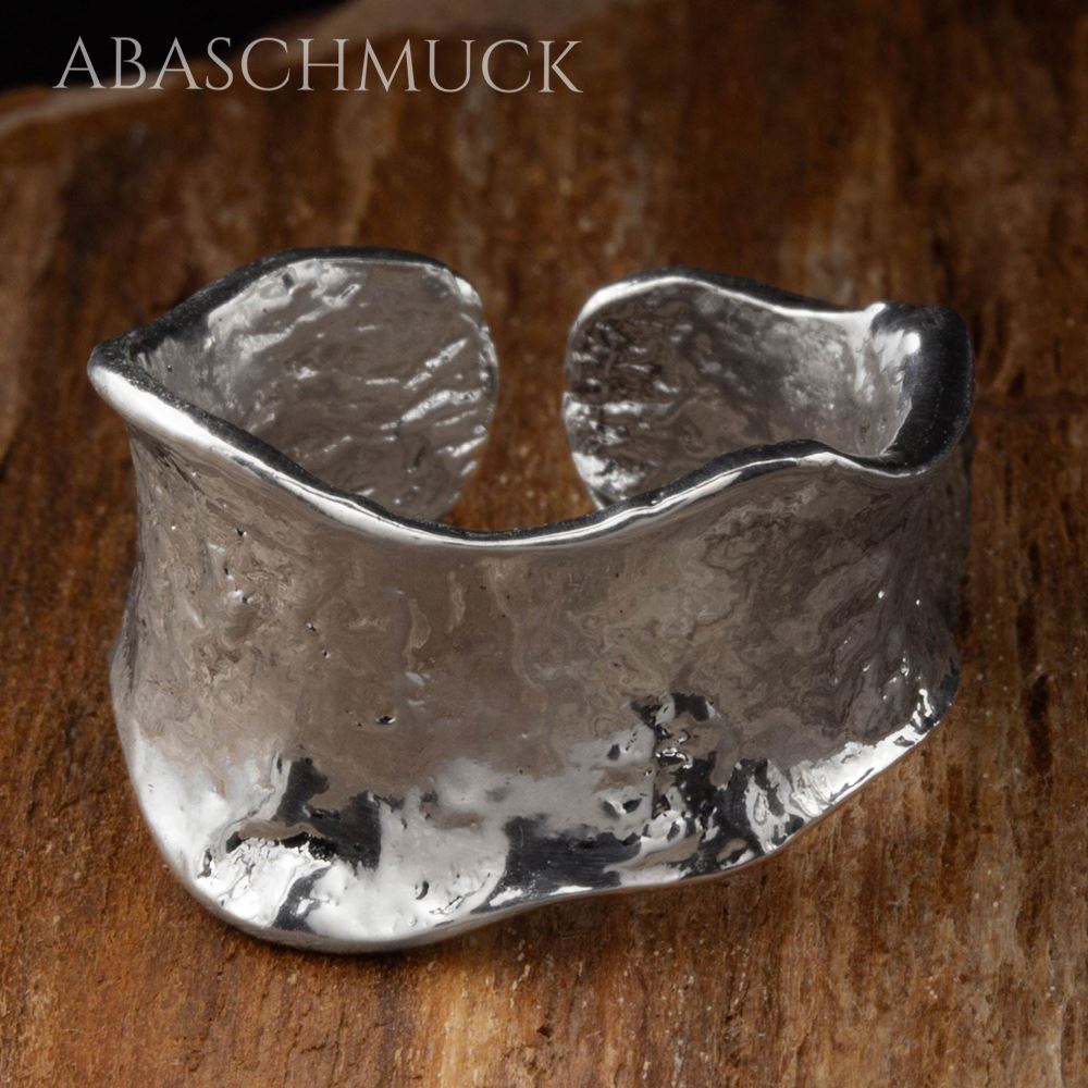 Breiter, welliger Silberring mit texturierter Oberfläche, natürliches Licht auf Holz.
